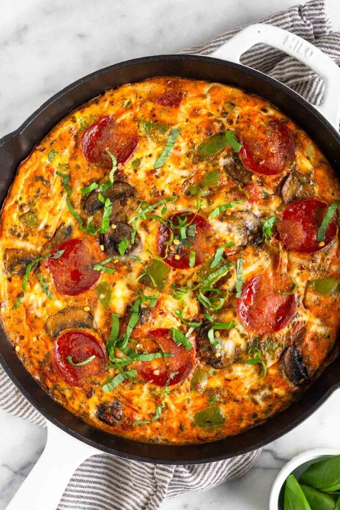 Pepperoni pizza frittata topped with fresh basil in a large white cast iron skillet. It is on a kitchen towel with a ramekin of basil next to it.
