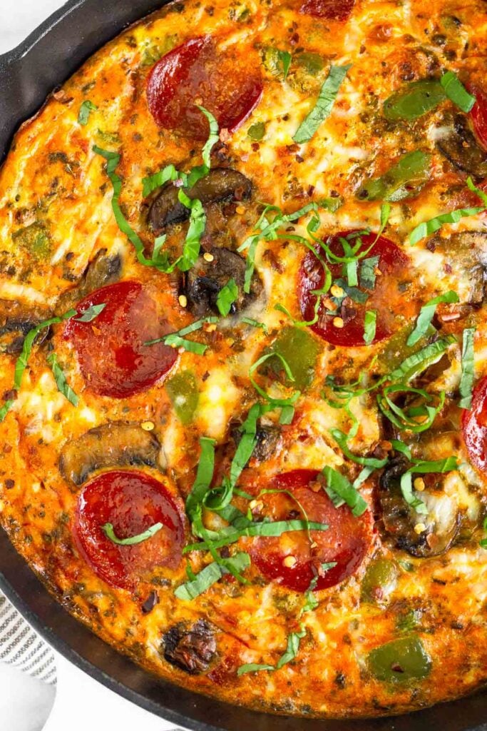 Close up of a pizza frittata with pepperoni topped with basil in a white skillet.
