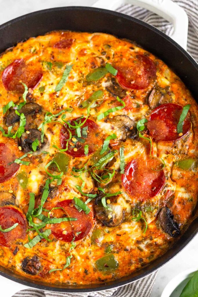 Breakfast pizza casserole with pepperoni and veggies in a large white skillet. It is topped with fresh basil and red pepper flakes.