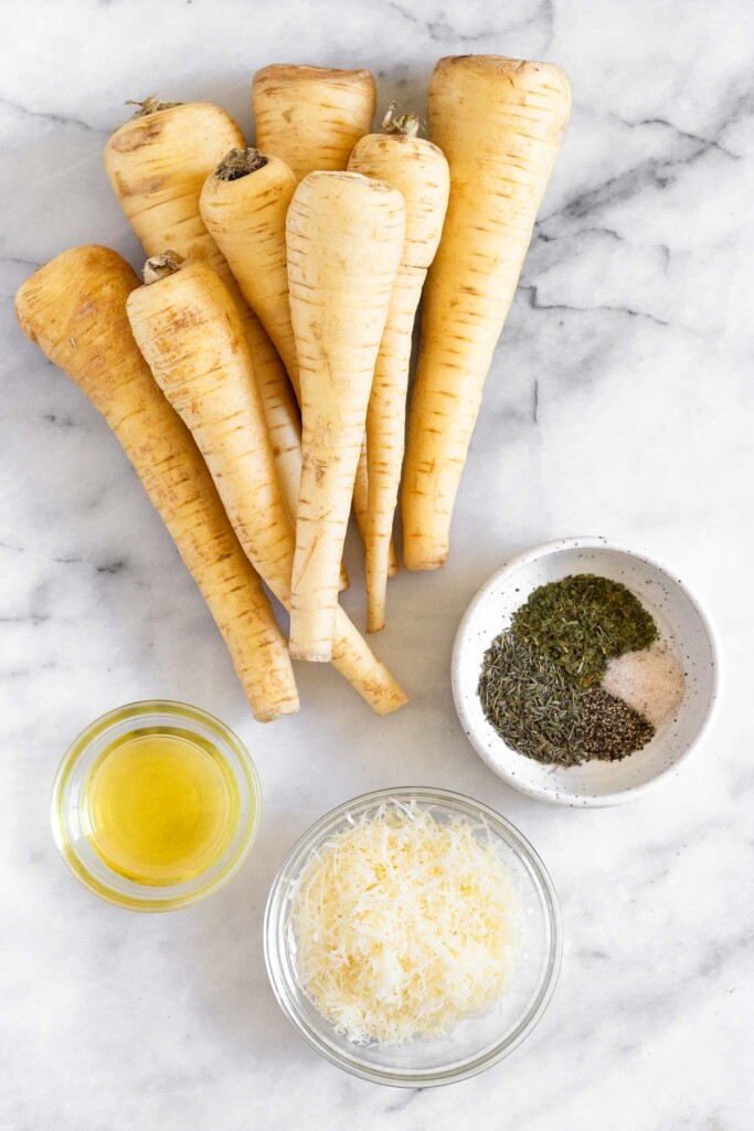White marble counter with a pile of parsnips, a bowl of spices, a bowl of grated parmesan cheese, and a bowl of olive oil.