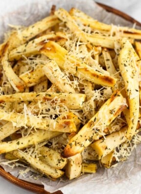 Roasted parsnips with crispy parmesan cheese and herbs on parchment paper on a wooden plate.