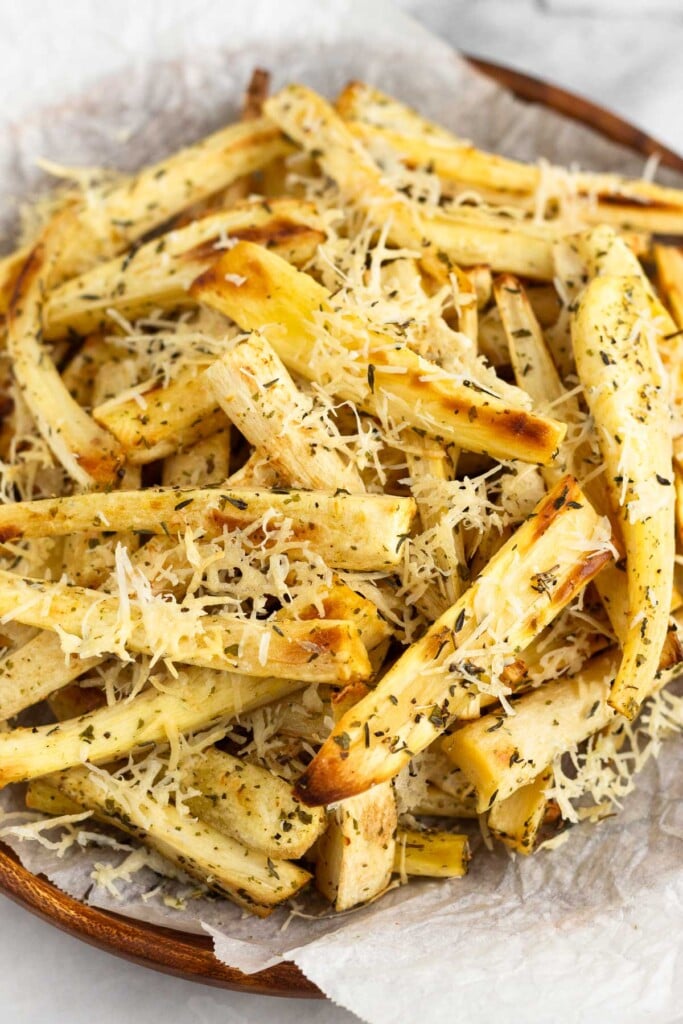 Roasted parsnips with crispy parmesan cheese and herbs on parchment paper on a wooden plate.