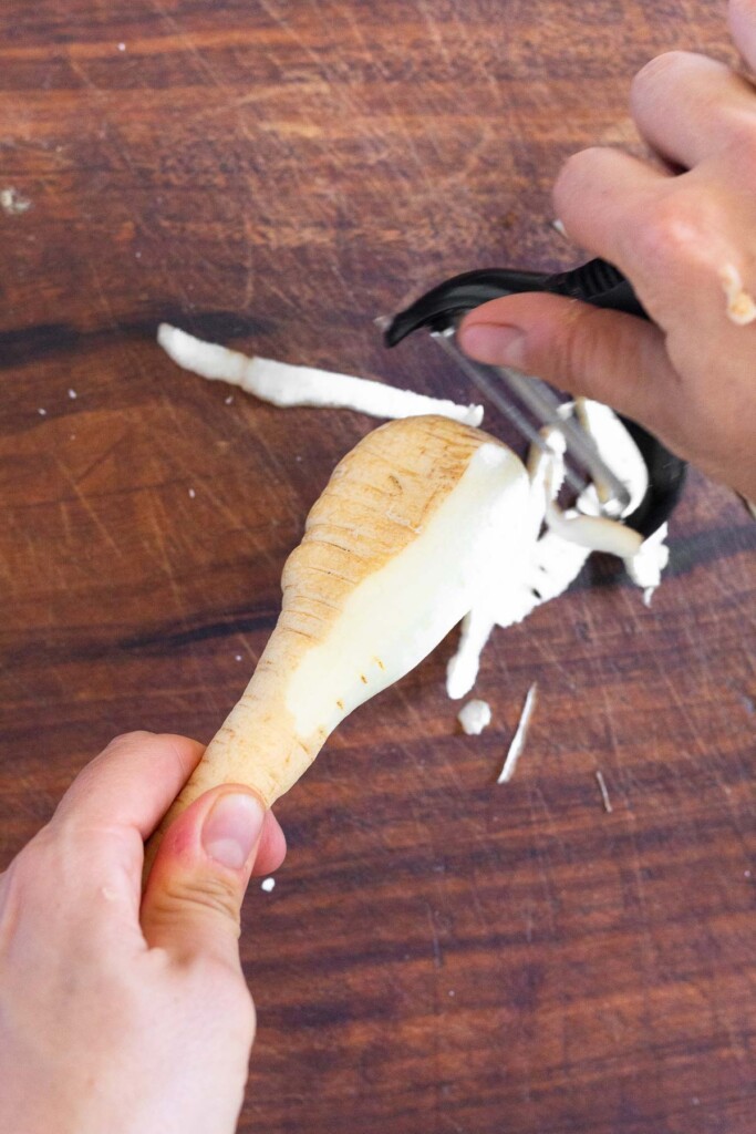 Overhead shot of someone peeling a parsnip over a wooden cutting board.