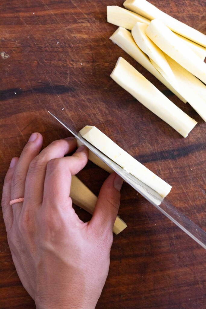 Overhead shot of someone cutting a peeled parsnip into matchsticks on wooden cutting board.