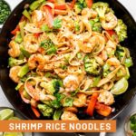 Shrimp stir fry with noodles in a large pan topped with red pepper flakes, cilantro, and green onion. Around it is a plate of lime wedges and small bowls of cilantro, green onions, and red pepper flakes.