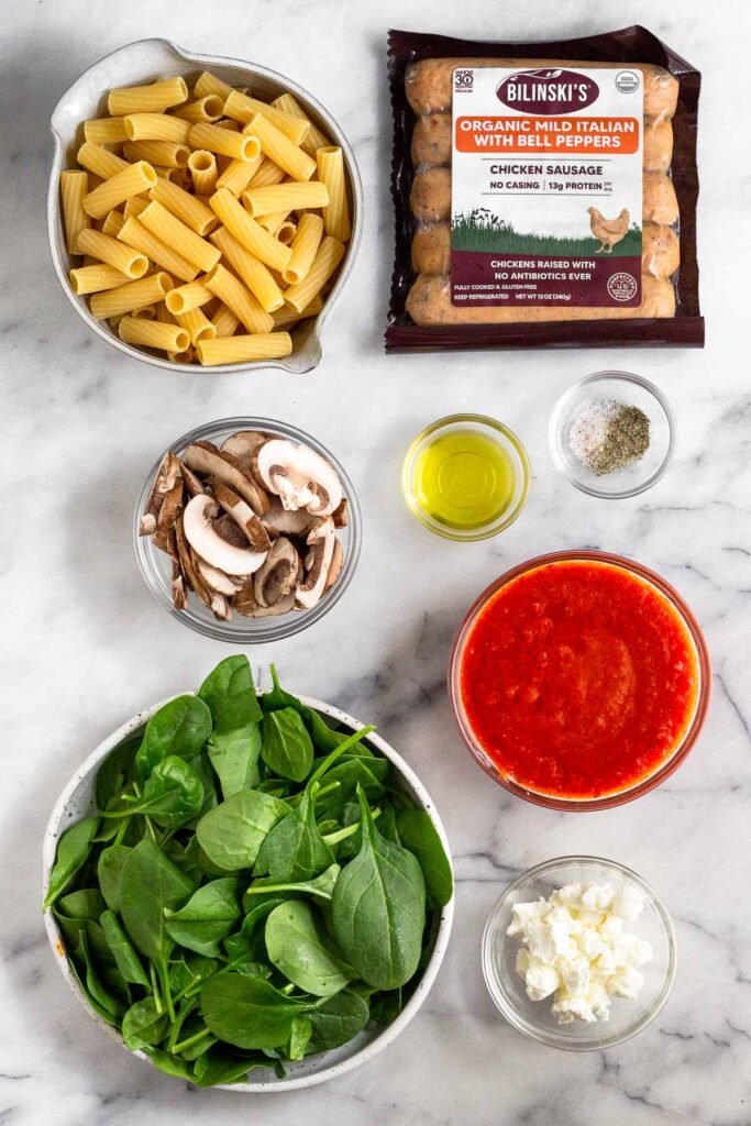 White counter with a bowl of uncooked rigatoni pasta, a package of chicken sausages, a bowl of salt and pepper, a bowl of olive oil, a bowl of marinara sauce, a bowl of goat cheese, a bowl of spinach, and a bowl of sliced mushrooms.