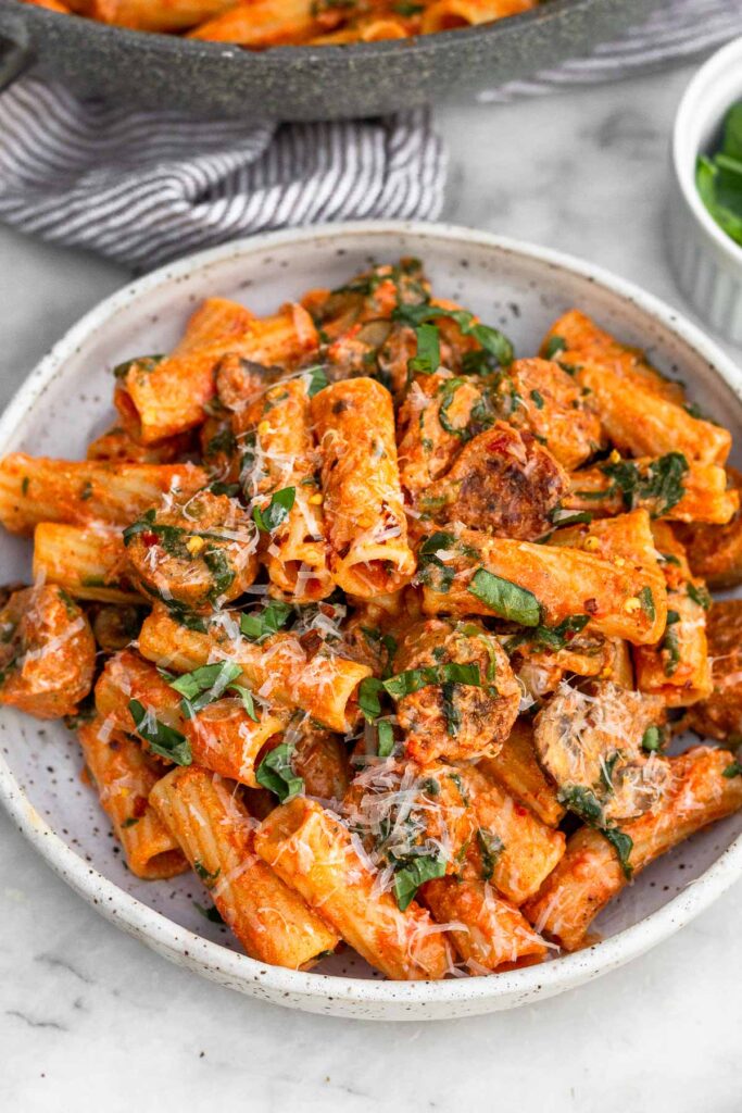 Chicken sausage with pasta, veggies, and marinara sauce on a white plate topped with basil and parmesan cheese. Next to the plate is a small ramekin of basil and a skillet with more pasta.