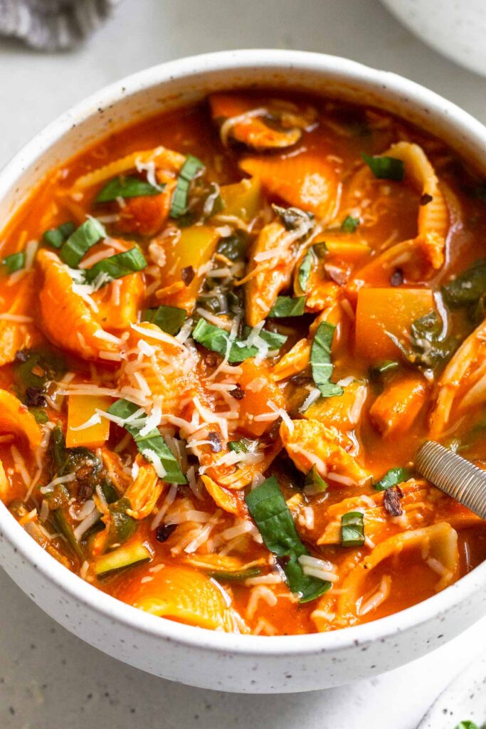 A bowl of chicken tomato soup with veggies and pasta. It is topped with fresh herbs and cheese with a spoon in it.