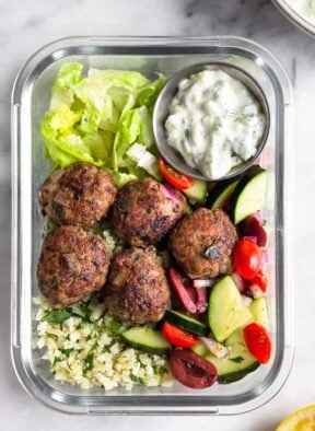 A high protein meal prep bowl filled with lamb meatballs, cucumber and tomato salad, cauliflower rice, chopped romaine lettuce, and a small dish of tzatziki.