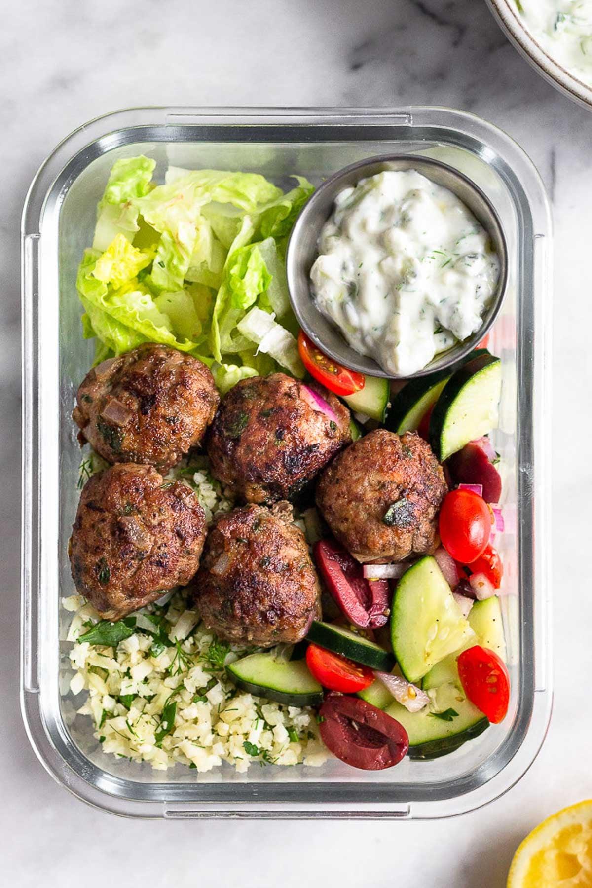 A high protein meal prep bowl filled with lamb meatballs, cucumber and tomato salad, cauliflower rice, chopped romaine lettuce, and a small dish of tzatziki.