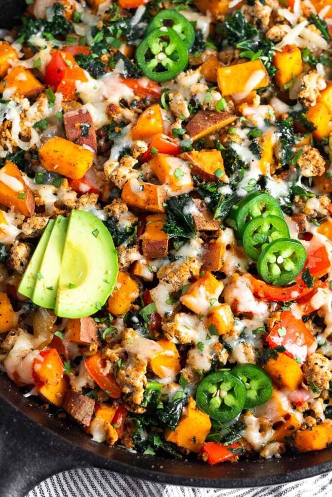Large cast iron filled with ground turkey, sweet potatoes, peppers, and kale, and topped with melted cheese. It is garnished with sliced avocado and jalapeños.