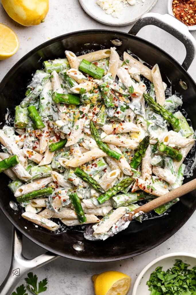 Pan filled with lemon asparagus pastas topped with red pepper flakes. Around it are some fresh lemons, a bowl of herbs, and a bowl of red pepper flakes.