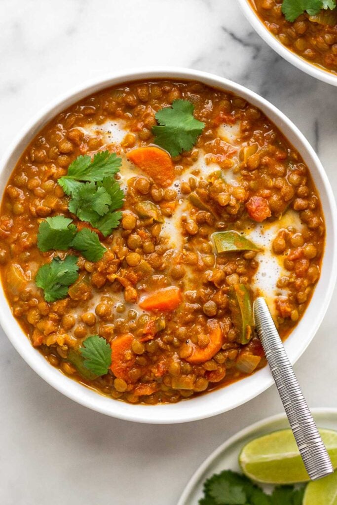 Bowl of lentil soup topped with a little coconut milk and cilantro with a spoon in it. Off to the side is another bowl of soup and a plate of limes and fresh herbs.