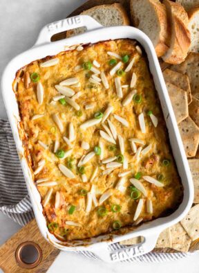 Hot crab dip in a small casserole dish topped with green onions and slivered almonds. Around is it sliced bread and crackers.