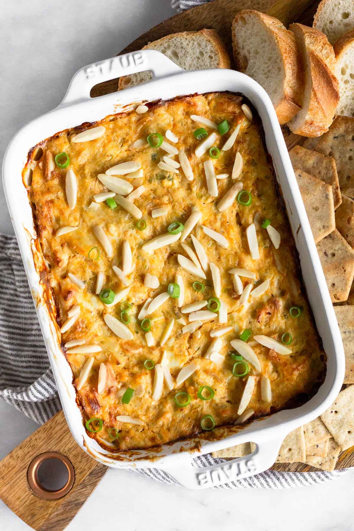 Hot crab dip in a small casserole dish topped with green onions and slivered almonds. Around is it sliced bread and crackers.