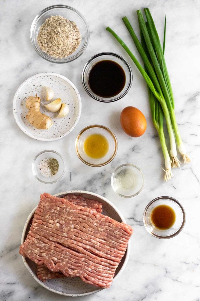 White counter top with a bowl of almond flour, a bowl of coconut aminos, a few green onions, an egg, a bowl of sesame oil, a bowl of rice vinegar, a bowl of fish sauce, a large bowl of ground pork, a bowl of salt and pepper, and small plate with fresh ginger and garlic cloves.
