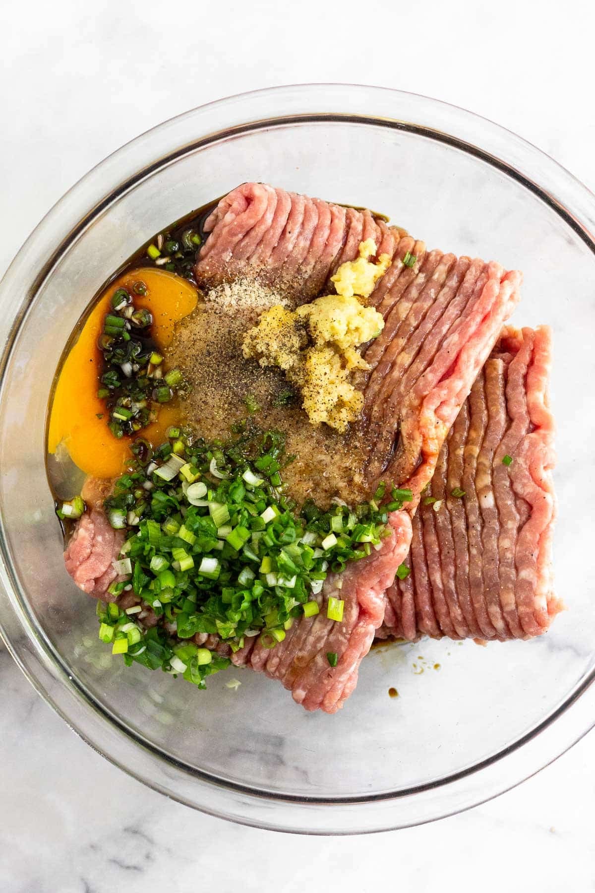 A large glass bowl with ground pork, an egg, minced scallions, garlic, ginger, coconut aminos, and spices.