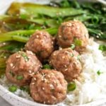 Pork meatballs on top on white rice with sautéed bok choy in a bowl. It is topped with sliced green onions and sesame seeds.