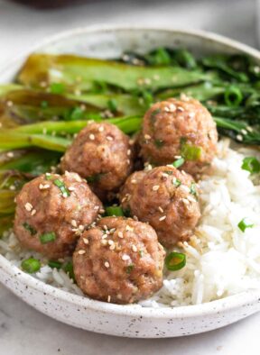 Pork meatballs on top on white rice with sautéed bok choy in a bowl. It is topped with sliced green onions and sesame seeds.