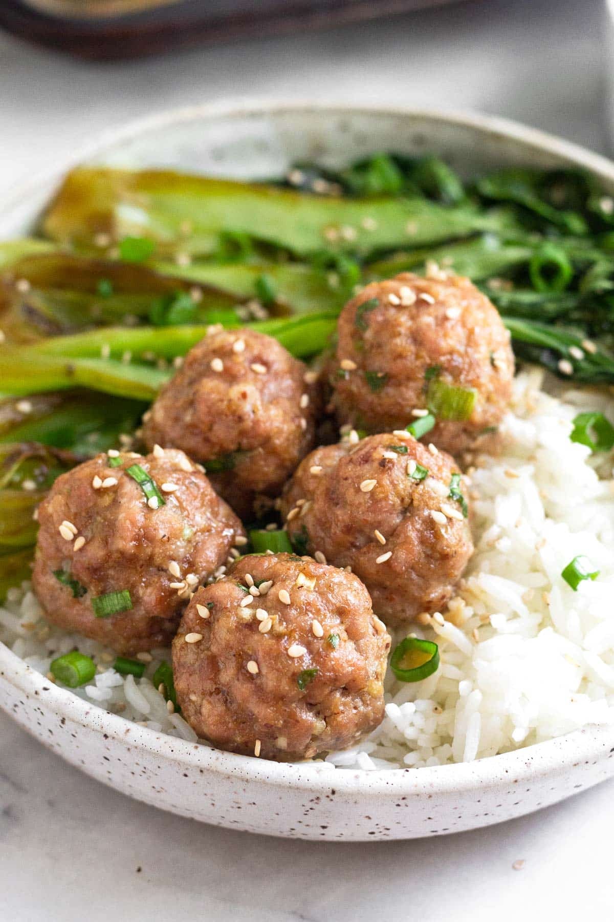 Pork meatballs on top on white rice with sautéed bok choy in a bowl. It is topped with sliced green onions and sesame seeds.