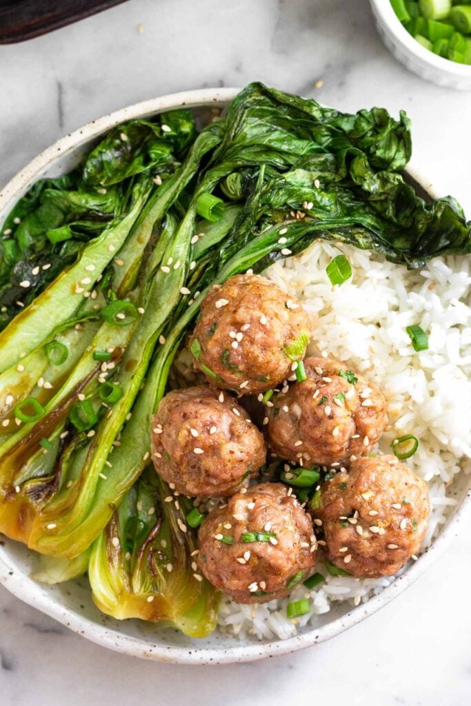 A bowl of white rice, sautéed bok choy, and asian pork meatballs. Everything is topped with green onions and sesame seeds.