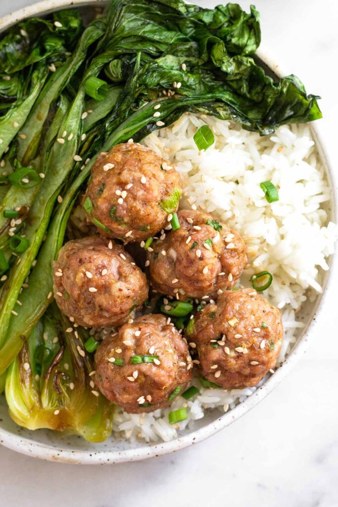 Baked pork meatballs in bowl with white rice and sautéed bok choy. Everything is garnished with sliced green onions and sesame seeds.