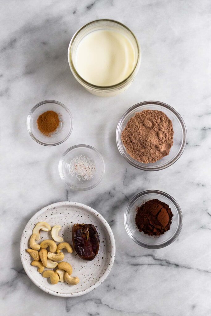 White marble counter with a jar of milk, a small bowl of chocolate protein powder, a small bowl of cocoa powder, a plate with some cashews and a medjool date on it, a small bowl of salt, and a small bowl of cinnamon.