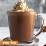A glass mug filled with protein hot chocolate topped with whipped cream and cinnamon. The mug is on a small saucer and behind it is a jar of milk.