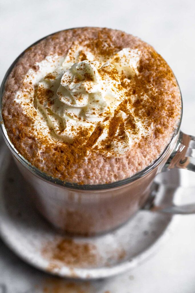 Overhead shot of protein powder hot chocolate in a glass mug topped with whipped cream and cinnamon. The mug is sitting on a small plate.