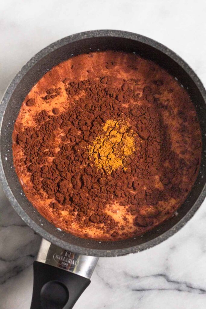 Saucepan filled with milk topped with cocoa powder and cinnamon before it is whisked together.