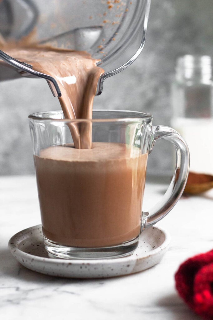 Healthy hot chocolate being poured into a glass mug from a blender. There is a red towel in front of the mug and a jar of milk behind the mug.