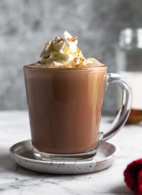 A glass mug filled with protein hot chocolate topped with whipped cream and cinnamon. The mug is on a small saucer and behind it is a jar of milk.