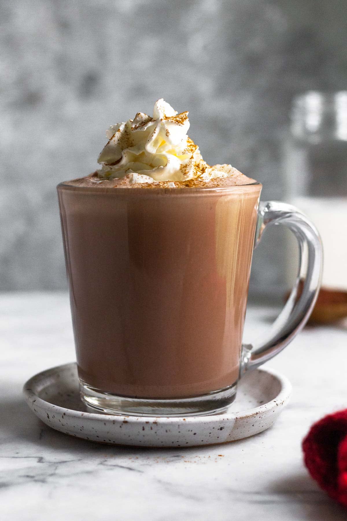A glass mug filled with protein hot chocolate topped with whipped cream and cinnamon. The mug is on a small saucer and behind it is a jar of milk.