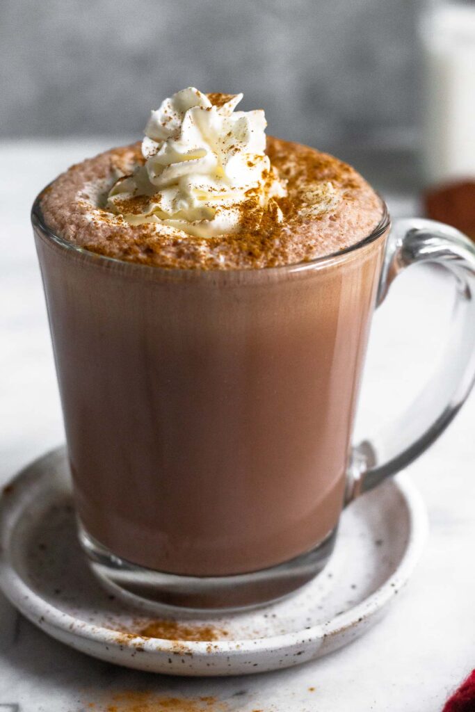 Large glass mug filled with healthy hot chocolate topped with whipped cream and cinnamon. The mug is sitting on a small plate and behind it is a jar of milk.