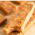 Two birthday cake protein bars stacked on top of each other with a bite taken out of the top one. Behind them are more bars.