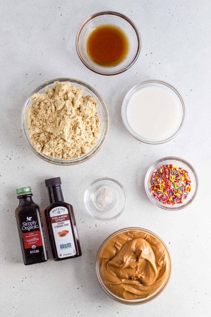 Overhead shot of a small bowl of maple syrup, a small bowl of milk, a small bowl of rainbow sprinkles, a bowl of cashew butter, a small bowl of salt, a bottle of almond extract, a bottle of vanilla extract, and bowl of protein powder.