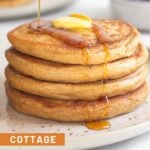 Cottage cheese pancakes with butter on them and syrup being poured on them. Behind the plate is a bowl of more berries and a plate with another stack of pancakes.