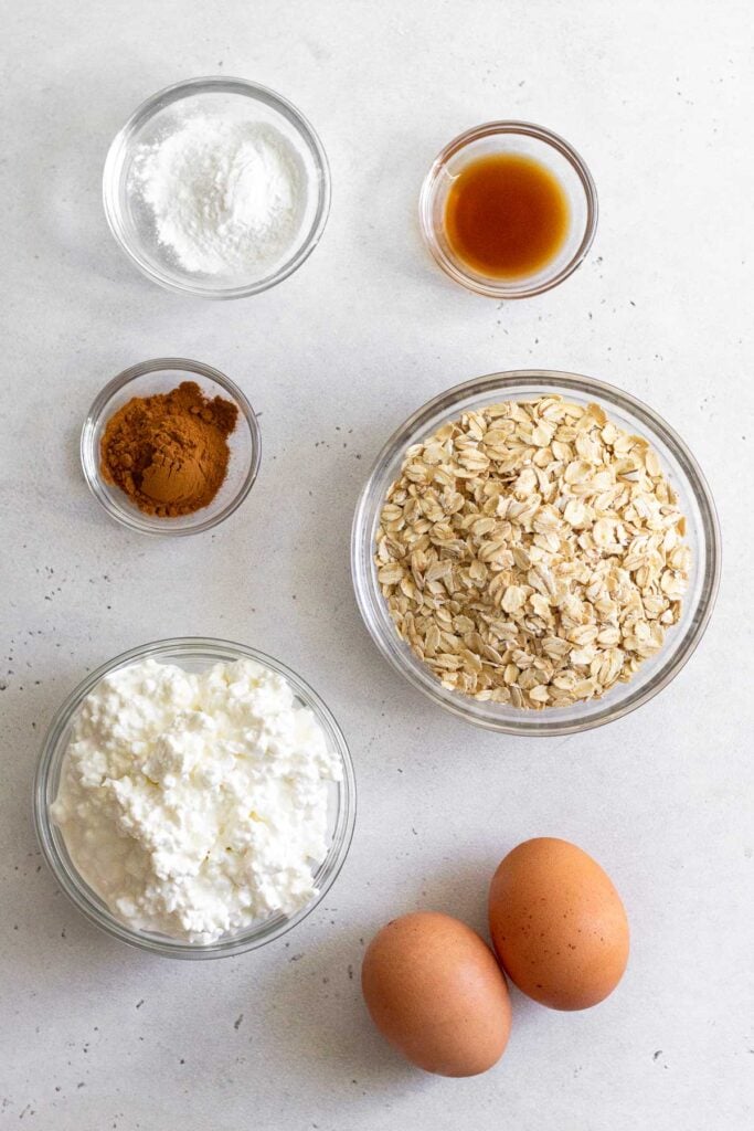 White counter with a bowl of baking powder, a bowl of vanilla extract, a bowl of rolled oats, 2 eggs, a bowl of cottage cheese, and a bowl of cinnamon.