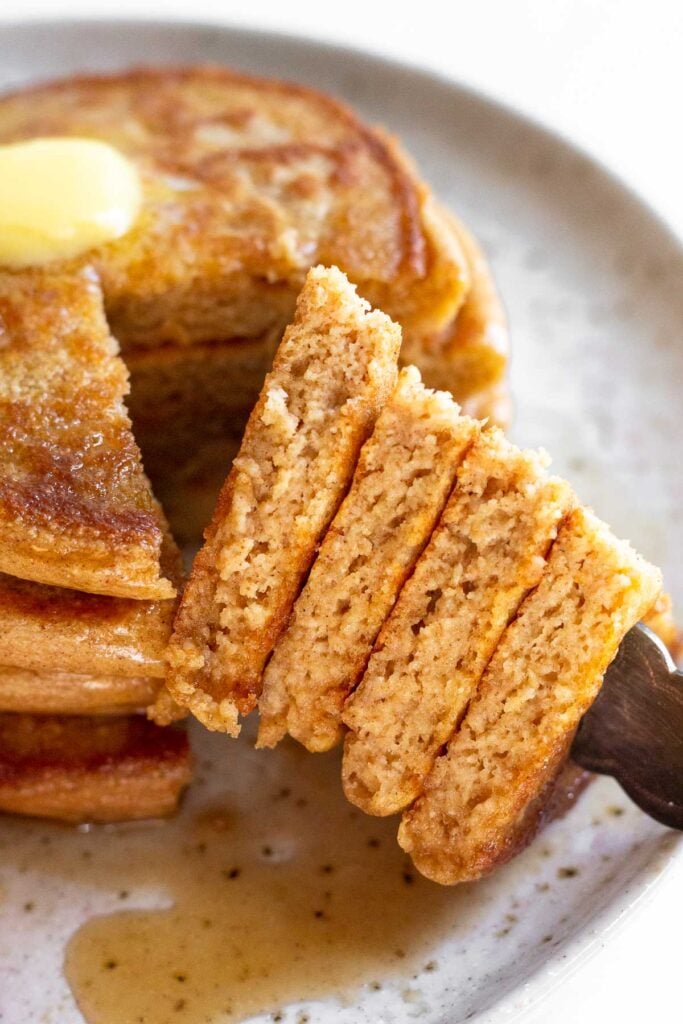 Close up of a stack of cottage cheese pancakes on a plate with a some cut out of them. There is a fork holding the cut pieces.