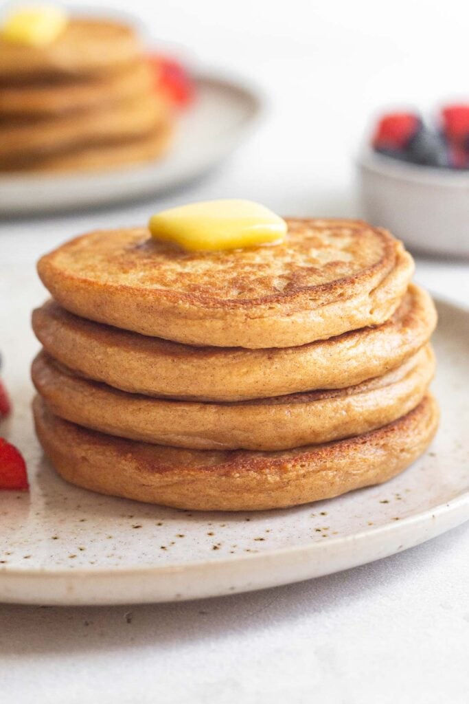 A stack of cottage cheese protein pancakes on a plate with butter on top of them. Behind them is a bowl of berries and another plate of pancakes.