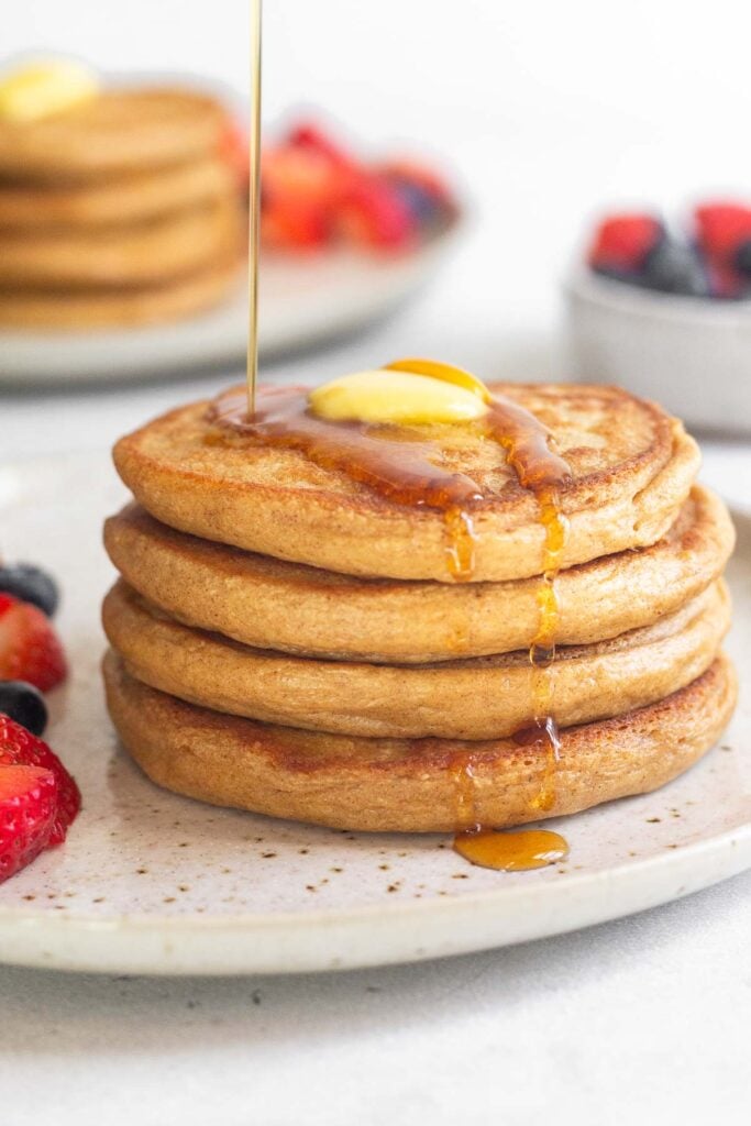 Oatmeal cottage cheese pancakes with butter on them and syrup being poured on them. Also on the plate are some berries. Behind the plate is a bowl of more berries and a plate with another stack of pancakes.