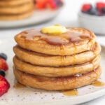 A stack of 4 cottage cheese pancakes with butter and syrup on a plate with fruit on it. Behind them is a bowl of more fruit and another stack of pancakes.