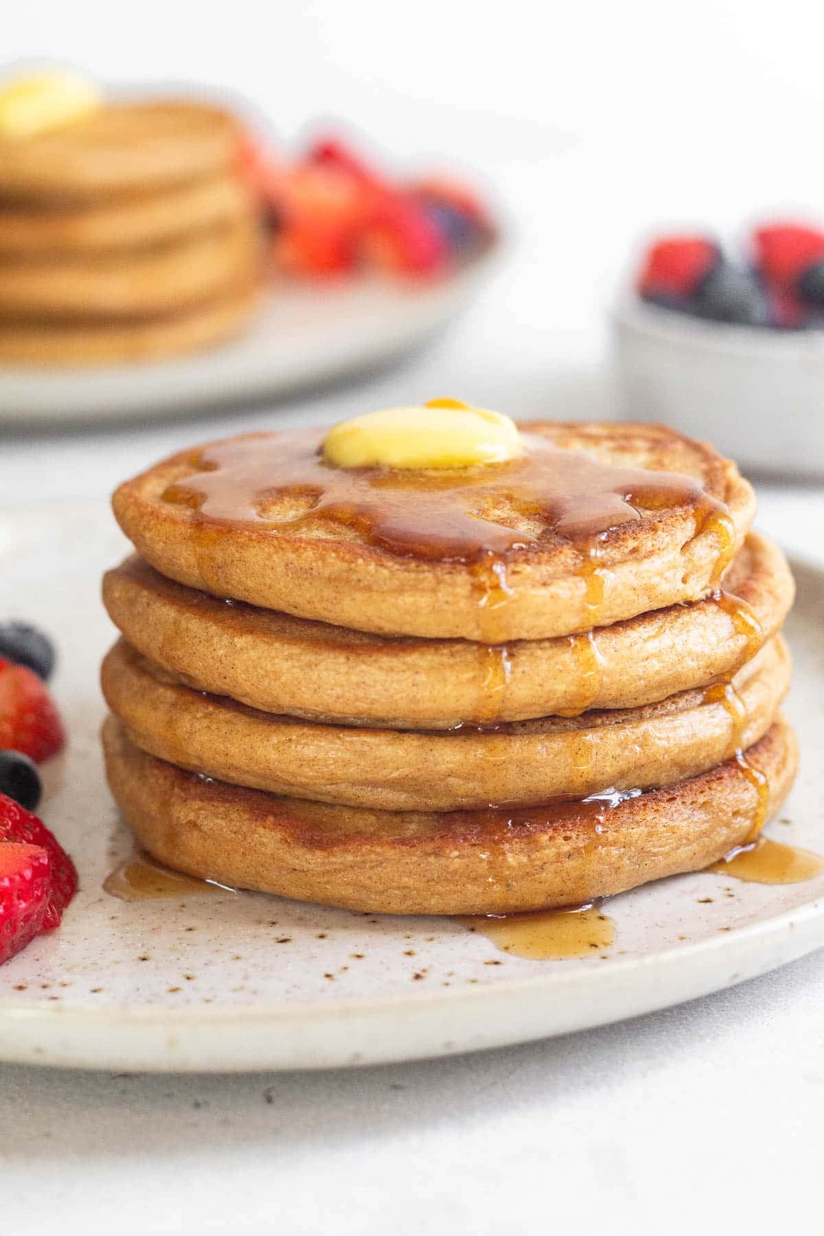A stack of 4 cottage cheese pancakes with butter and syrup on a plate with fruit on it. Behind them is a bowl of more fruit and another stack of pancakes.