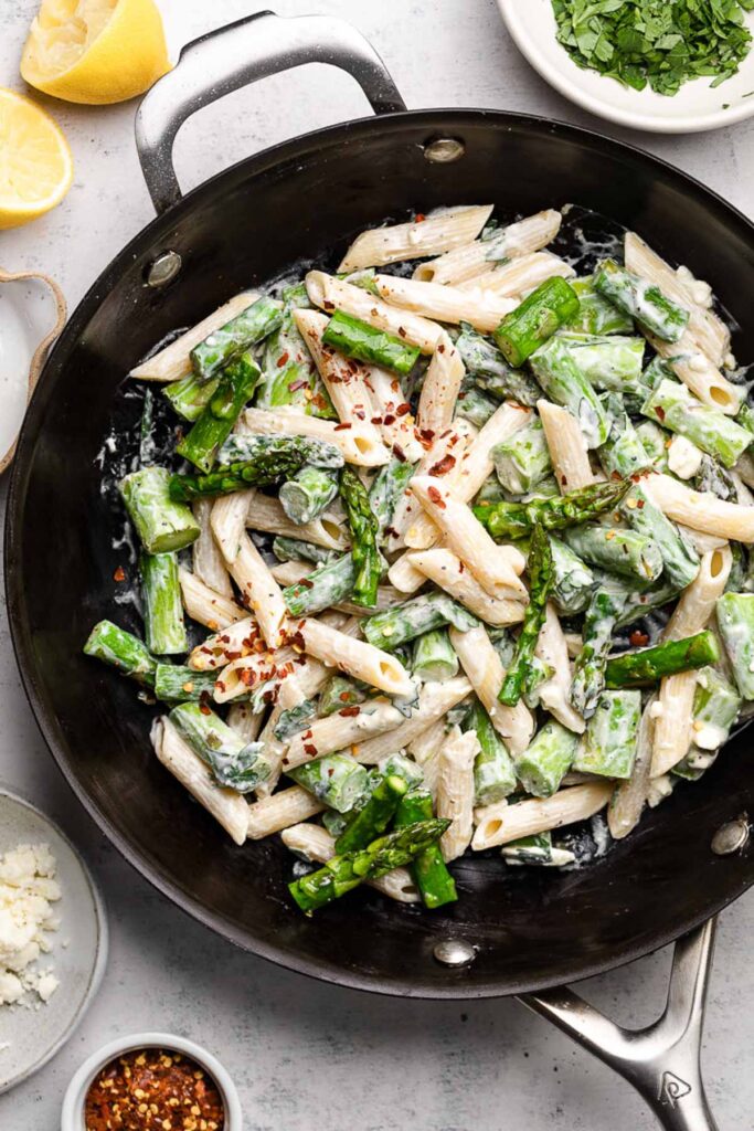 Skillet filled with pasta and asparagus mixed with a white sauce. Around it is bowl of spices, a bowl of crumbled cheese, halves of lemons, and a bowl of herbs.