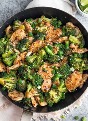 Macro friendly recipe with chicken and broccoli tossed in a brown sauce and garnished with green onions and sesame seeds in a large skillet. Next to it is a bowl of lime wedges.
