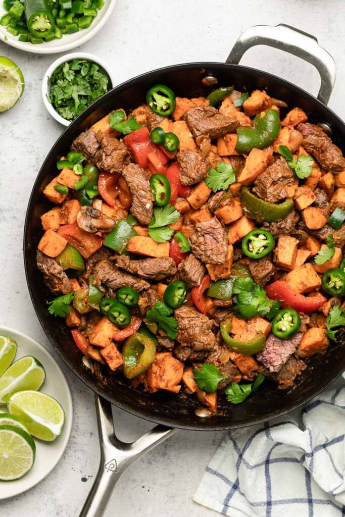 Macro friendly dinner recipe of steak, sweet potatoes, and peppers in a large skillet. Around it is a bowl of sliced jalapeños, a bowl of cilantro, and a bowl of lime wedges.