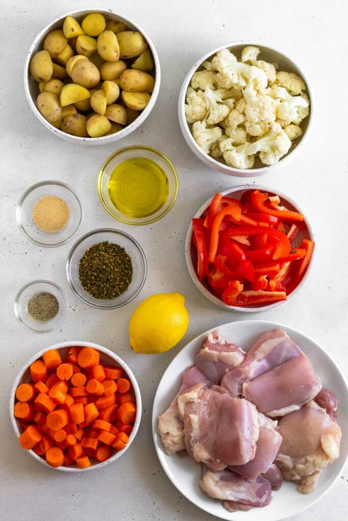 Overhead shot of a bowl of diced potatoes, a bowl of cauliflower florets, a bowl of julienned peppers, a plate of raw chicken thighs, a bowl of diced carrots, a lemon, three bowls of different spices, and a bowl of olive oil.