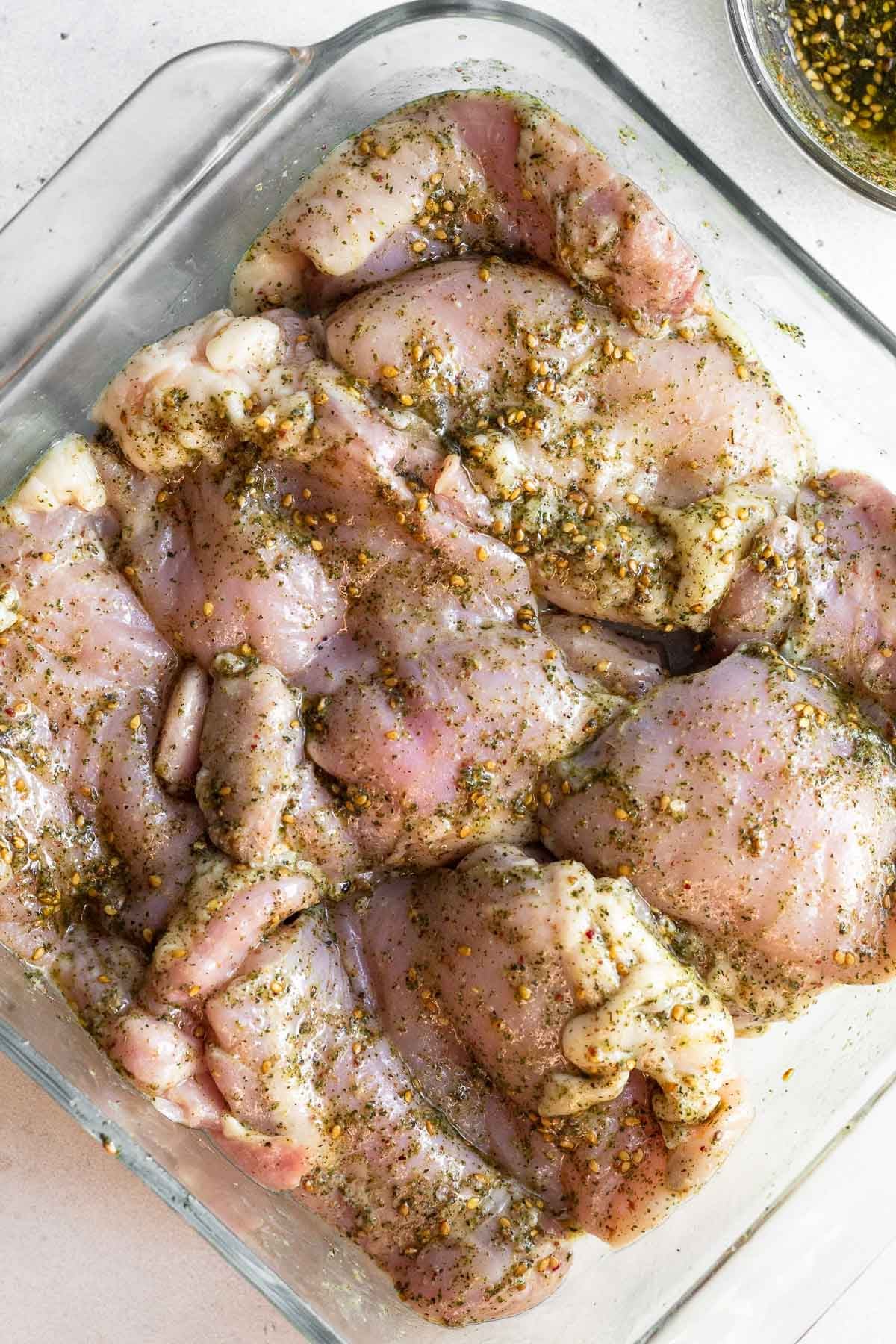 Raw chicken thighs in a baking dish covered with oil and za'atar seasoning.