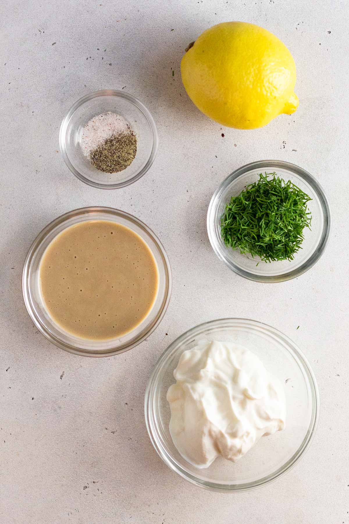 Overhead shot of a lemon, a bowl of fresh dill, a bowl of greek yogurt, a bowl of tahini, and a bowl of salt and pepper.