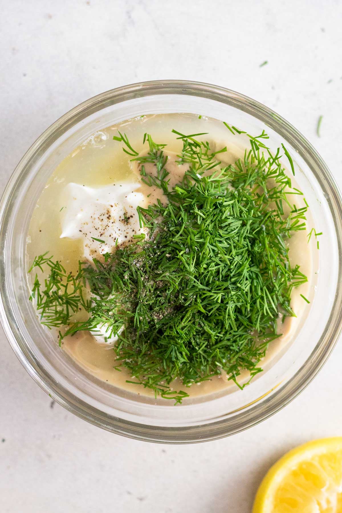 Small bowl with yogurt, tahini, lemon juice, and dill in it before it is mixed up. Next to it is half a lemon.